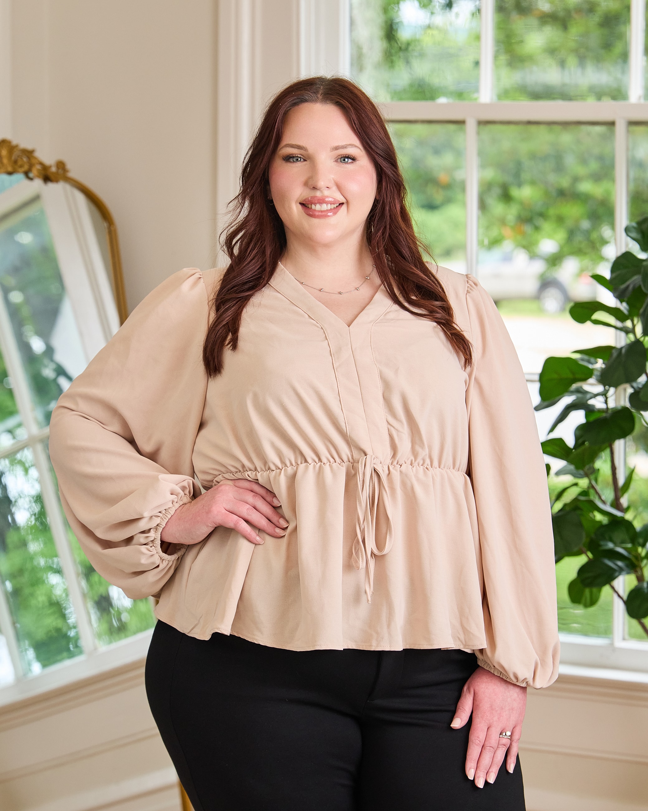 Smiling woman in blush top by windowsill.